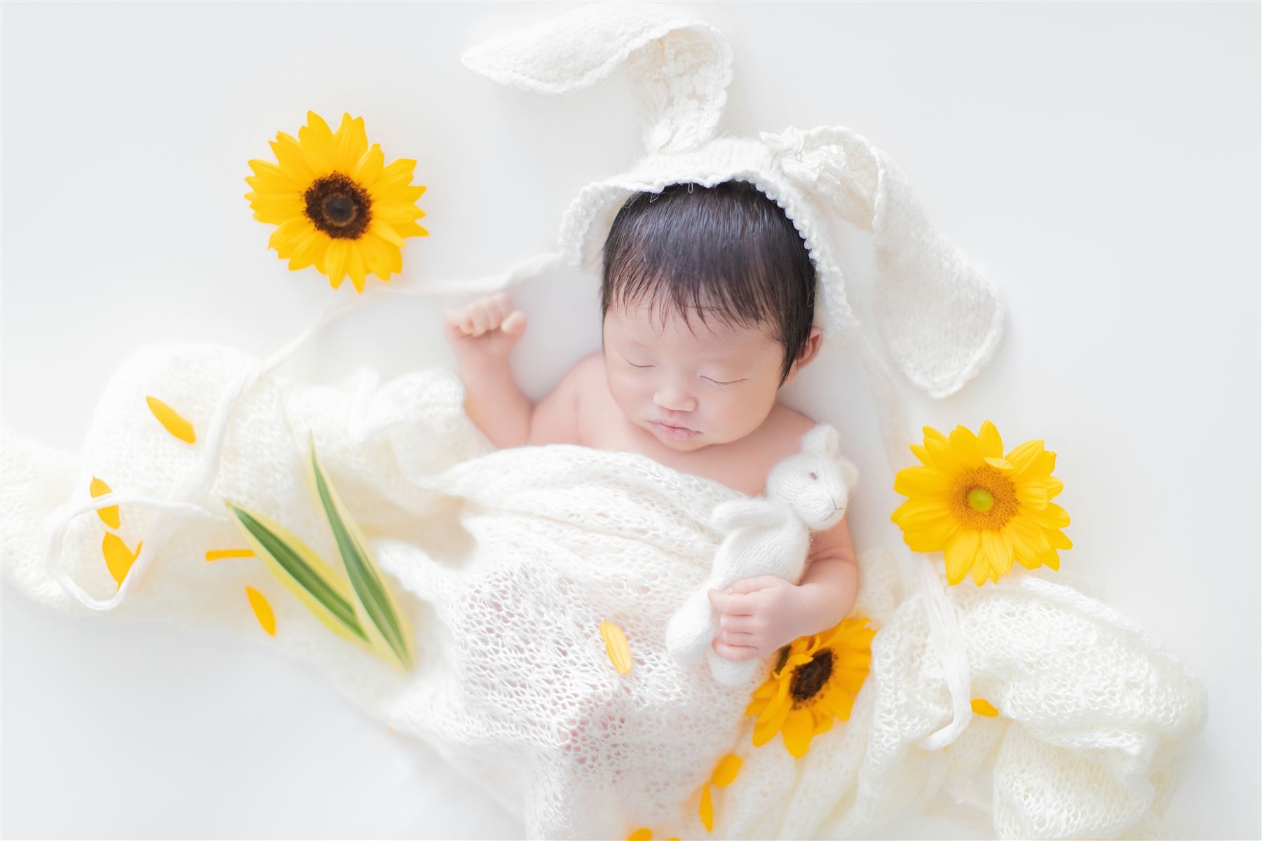 ひまわりに囲まれ、うさぎ耳のフードをかぶって眠る新生児
