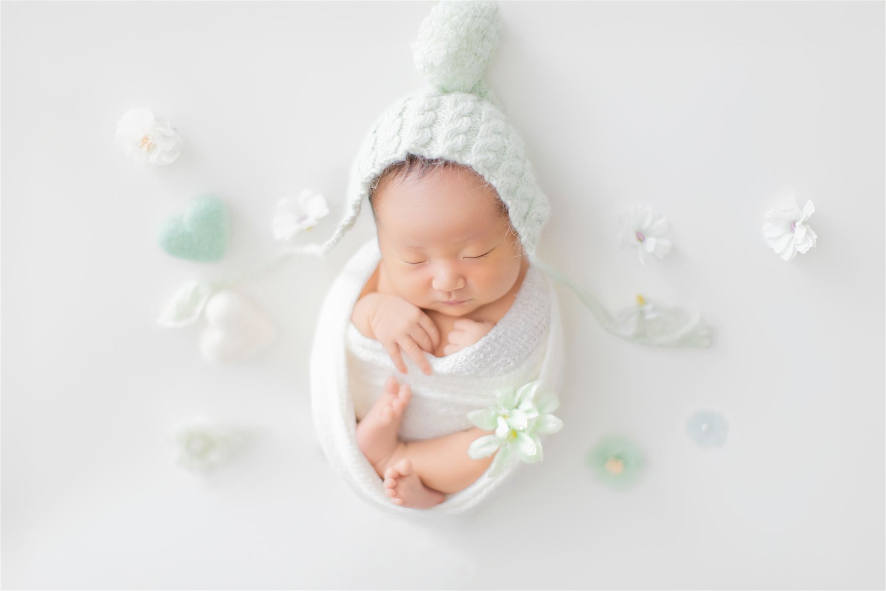 ミントグリーンの帽子をかぶり、白い花に囲まれて眠る新生児